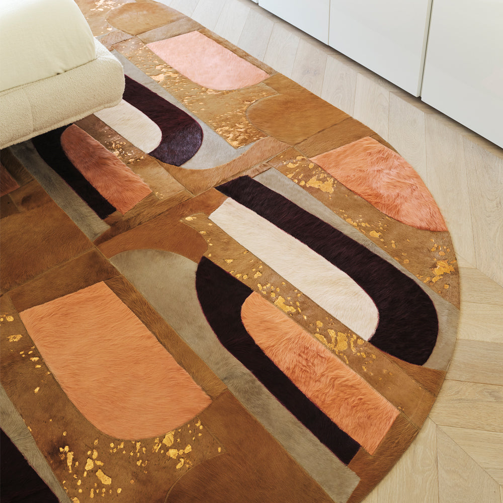 round cowhide rug featuring a patchwork of abstract, curved shapes in tan, burnt orange, and deep plum, accented with gold metallic flecks on a light wood floor.