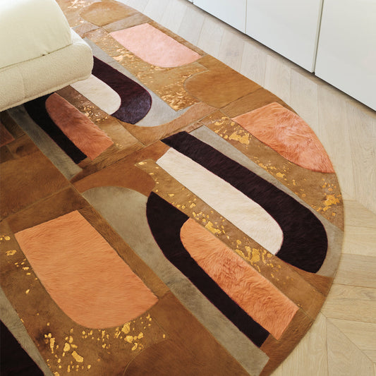 round cowhide rug featuring a patchwork of abstract, curved shapes in tan, burnt orange, and deep plum, accented with gold metallic flecks on a light wood floor.