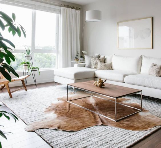 A bright, airy living room where a natural brown and white cowhide rug is layered over a larger textured area rug, placed beneath a minimalist wooden coffee table.
