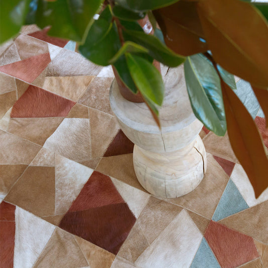 A close-up of a geometric cowhide rug with earthy brown, tan, and muted teal patches, styled with a leafy green plant.