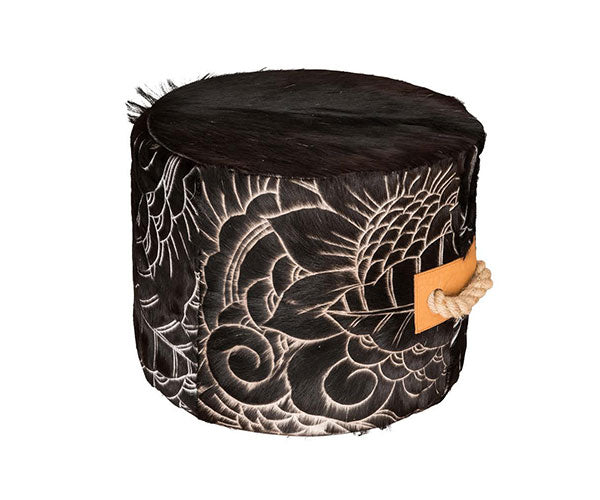 round cowhide ottoman with intricate, etched floral and swirl pattern in black and cream. Tan leather side tabs with thick, natural rope handles.