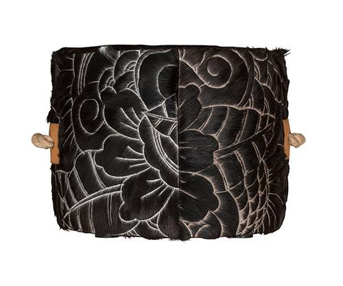 round cowhide ottoman with intricate, etched floral and swirl pattern in black and cream. Tan leather side tabs with thick, natural rope handles.