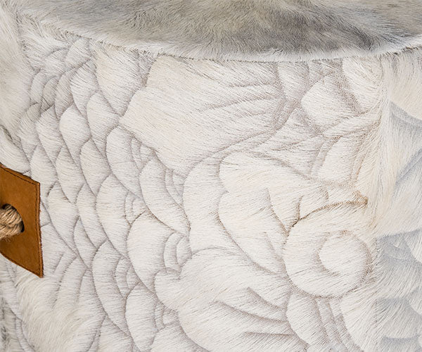 cowhide ottoman with intricate, etched floral and swirl pattern in gray and cream. Tan leather side tabs with thick, jute handles.