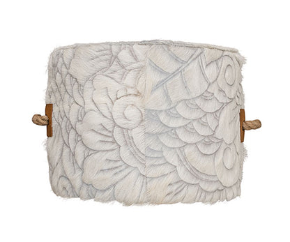 cowhide ottoman with intricate, etched floral and swirl pattern in gray and cream. Tan leather side tabs with thick, jute handles.